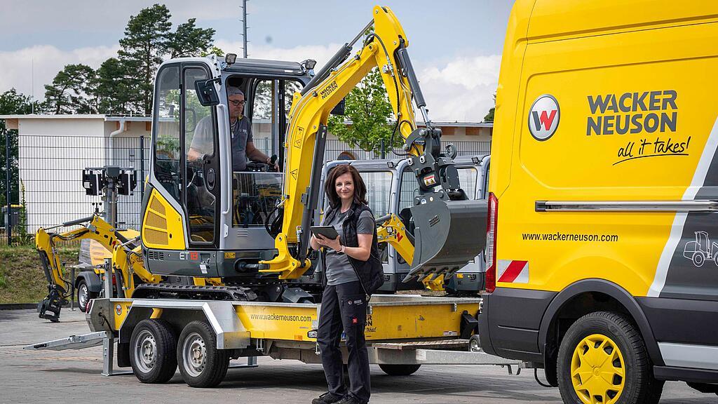 Bagger wird auf einen Hänger geladen, Mitarbeiterin steht mit Tablet davor. Servicemobil von Wacker Neuson im Hintergrund. 