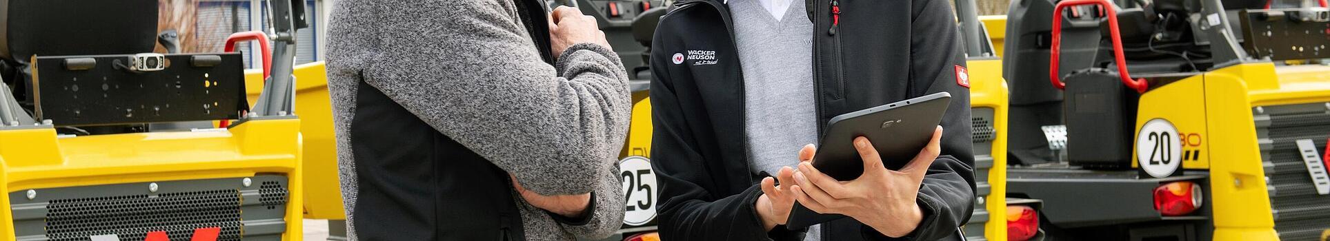 Lächelnde Wacker Neuson Mitarbeiterin mit Tablet berät lächelnden Kunden vor Wacker Neuson Baumaschinen.