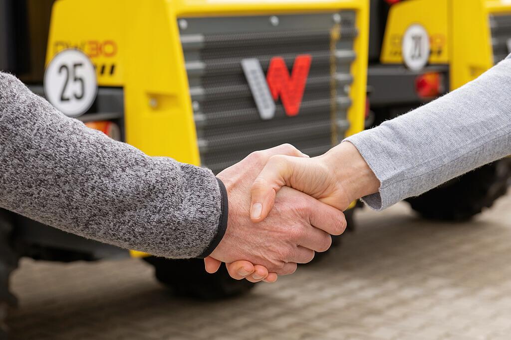Handschlag zwischen Wacker Neuson Mitarbeiterin und Kunde, Wacker Neuson Baumaschinen im Hintergrund.