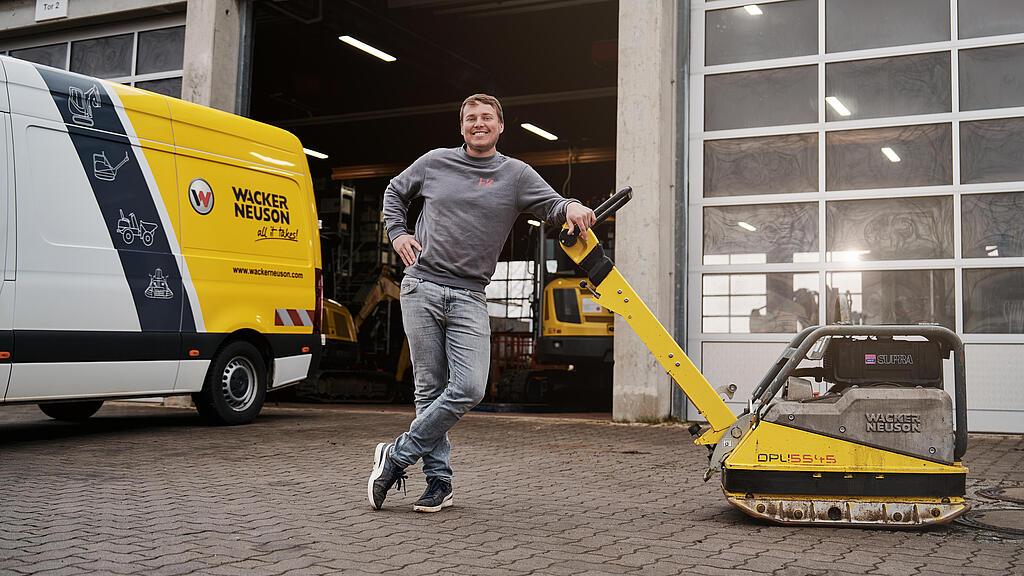 Ein Mann steht vor einer Werkstatt neben einer Rüttelplatte und einem Servicefahrzeug von Wacker Neuson. Im Hintergrund sind geöffnete Werkstatttore und weitere Baumaschinen zu sehen.