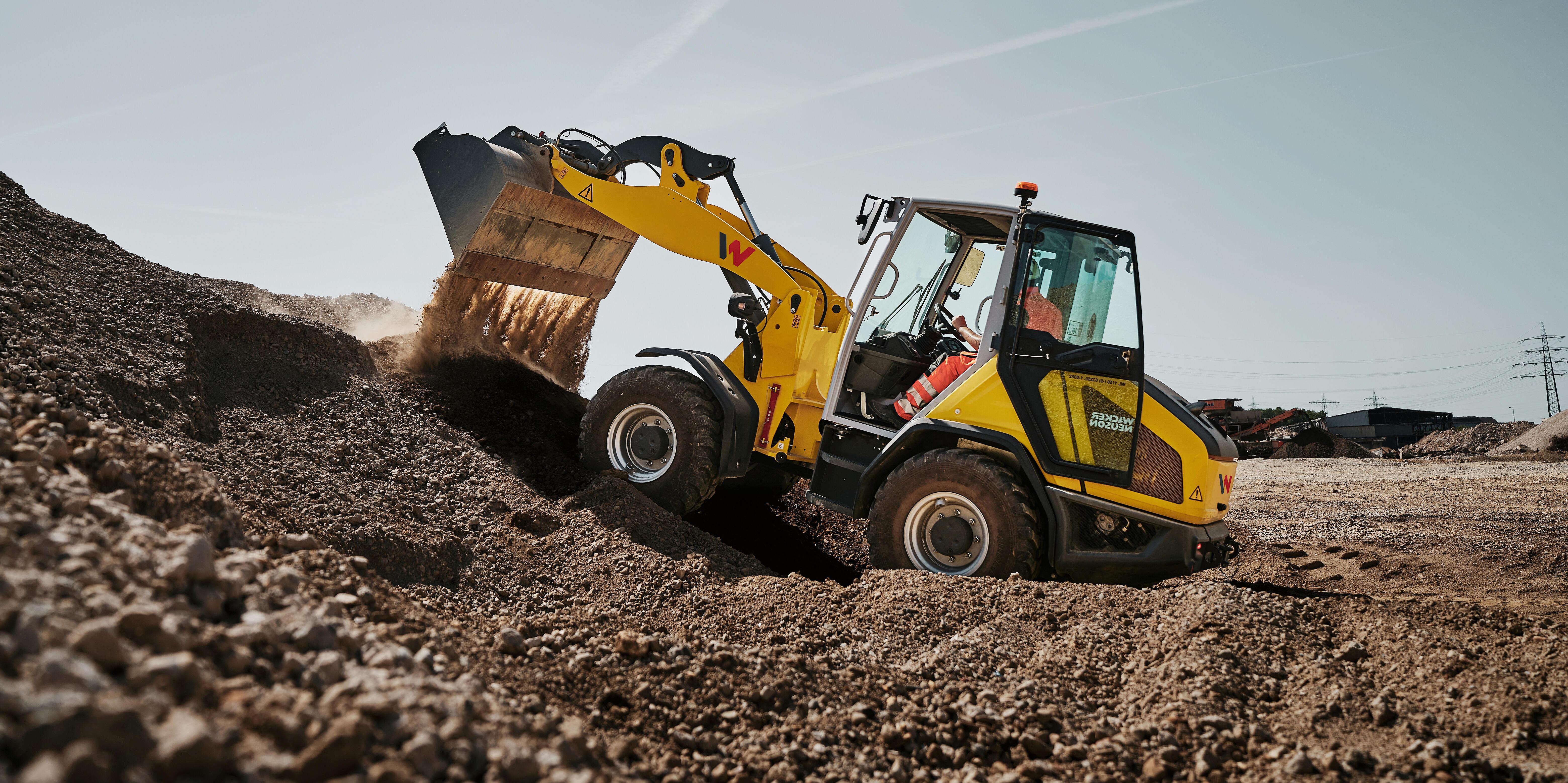 Wacker Neuson WL1150 wheel loader loading and moving aggregate material at a quarry site, with the bucket raised and material flowing from the bucket.