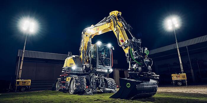 Wacker Neuson EW135 wheeled excavator equipped with a tiltrotator attachment, illuminated by floodlights on a construction site at night.