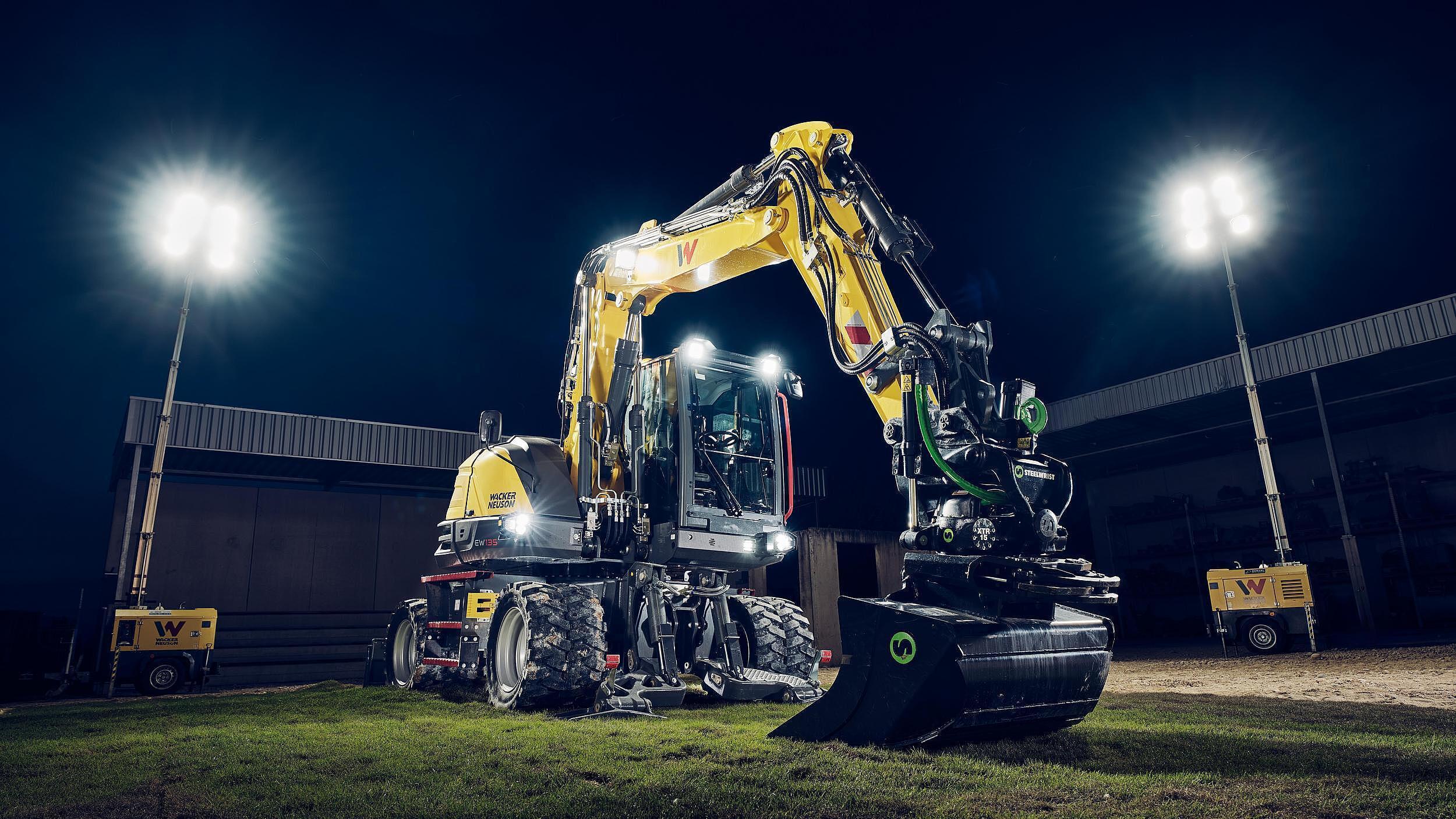 Wacker Neuson EW135 wheeled excavator equipped with a tiltrotator attachment, illuminated by floodlights on a construction site at night.