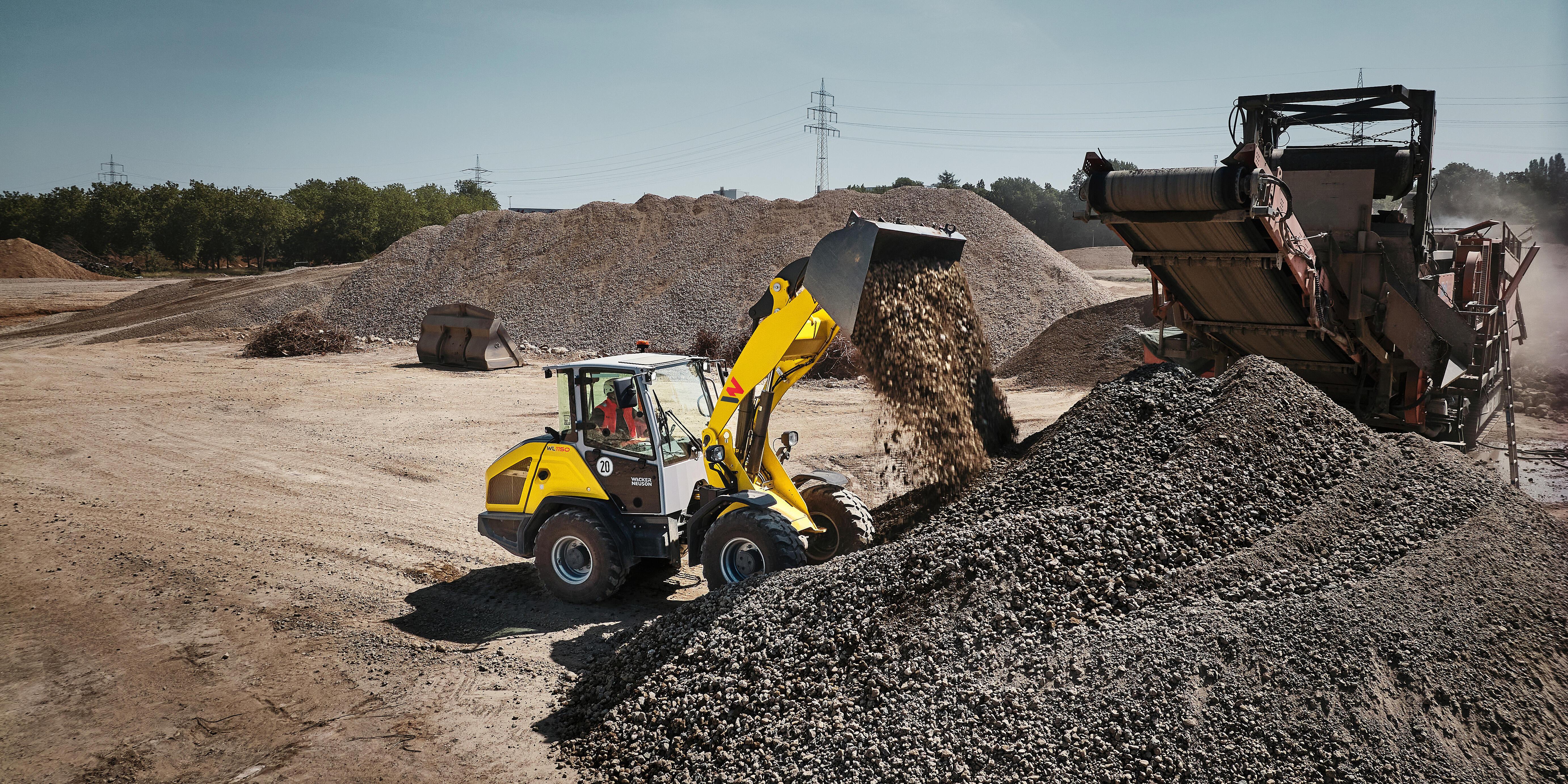 Wacker Neuson WL1150 wheel loader unloading gravel onto a stockpile at a quarry, with screening equipment and aggregate piles in the background.