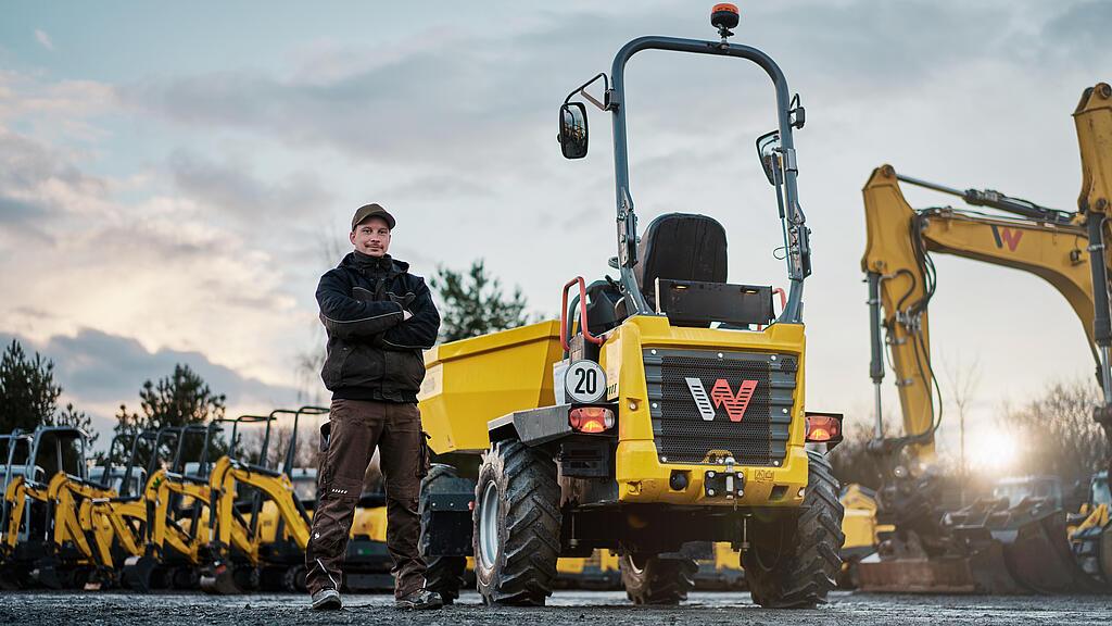 Ein Mann steht vor einem gelben Dumper von Wacker Neuson. Im Hintergrund stehen mehrere Minibagger und ein großer Bagger auf einem Baumaschinenhof.