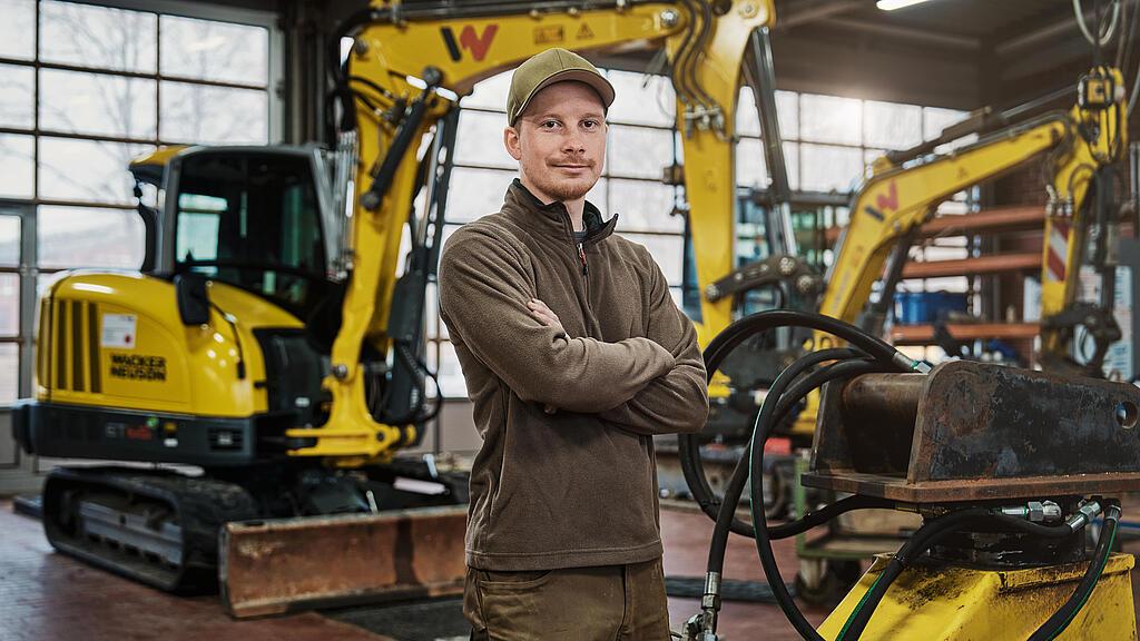 Ein Mann in Arbeitskleidung steht in einer Werkstatt vor mehreren gelben Minibaggern von Wacker Neuson. Im Vordergrund ist ein hydraulisches Anbaugerät zu sehen.