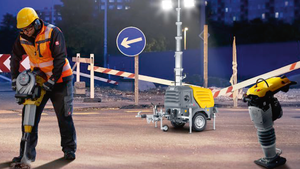A worker on a night construction site uses a Wacker Neuson demolition hammer; next to it are a light mast and a tamper.