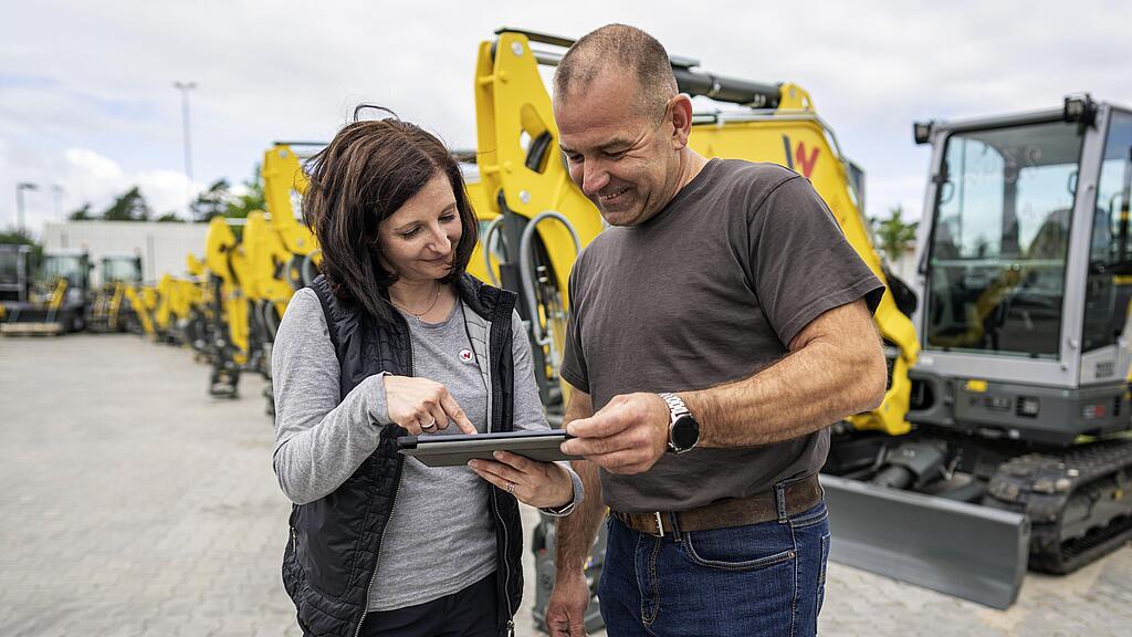 Wacker Neuson Mitarbeiter schauen gemeinsam in ein tablet, im Hintergrund sind viele Minibagger zu sehen.