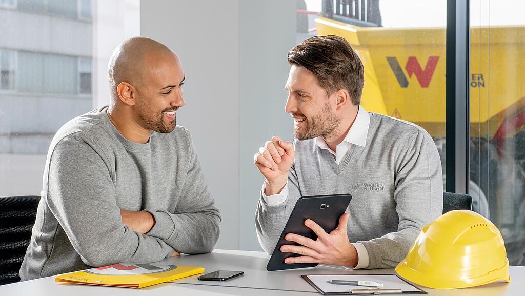 Wacker Neuson Mitarbeiter sitzt am Schreibtisch und zeigt Kunden etwas auf dem Tablet.