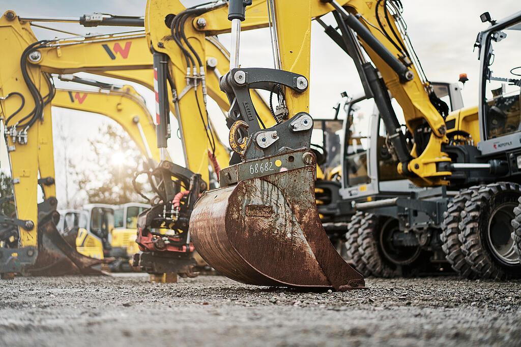 Several yellow excavators are lined up on a gravel yard. A large, slightly rusty excavator bucket is clearly visible in the foreground. The machines are clean and appear to be in good condition, ready for sale or use. The sun is shining through the trees in the background, giving the image a warm, bright atmosphere.