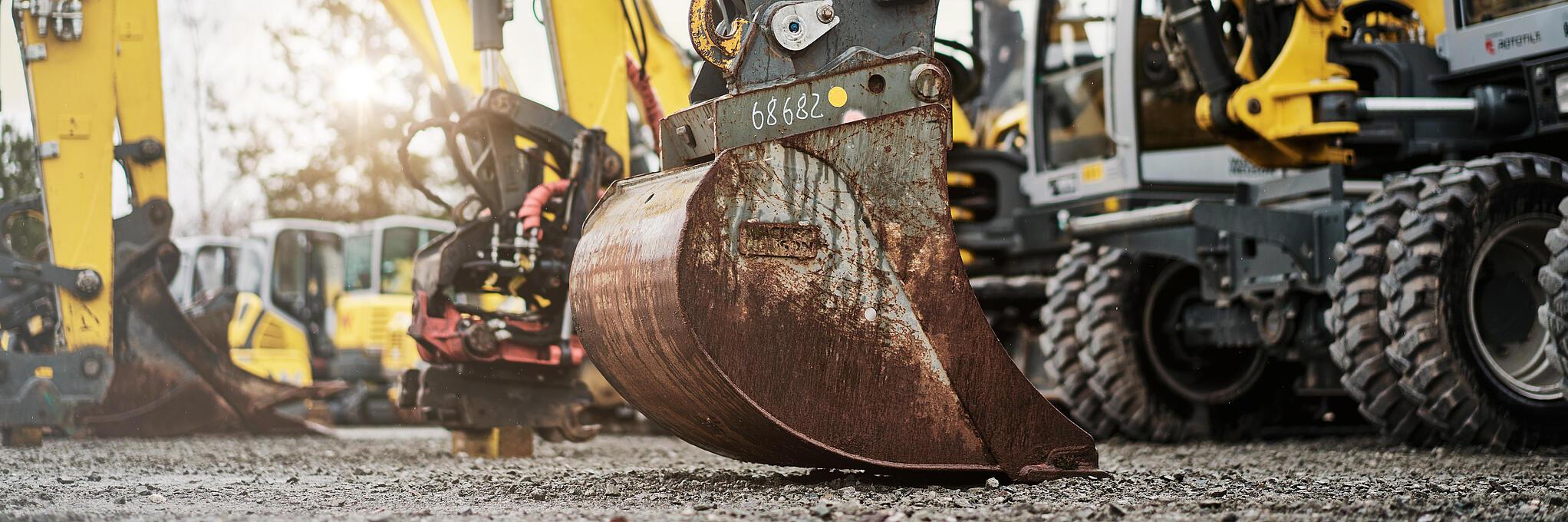 Several yellow excavators with hydraulic arms on a gravel site, in the foreground a worn excavator shovel.