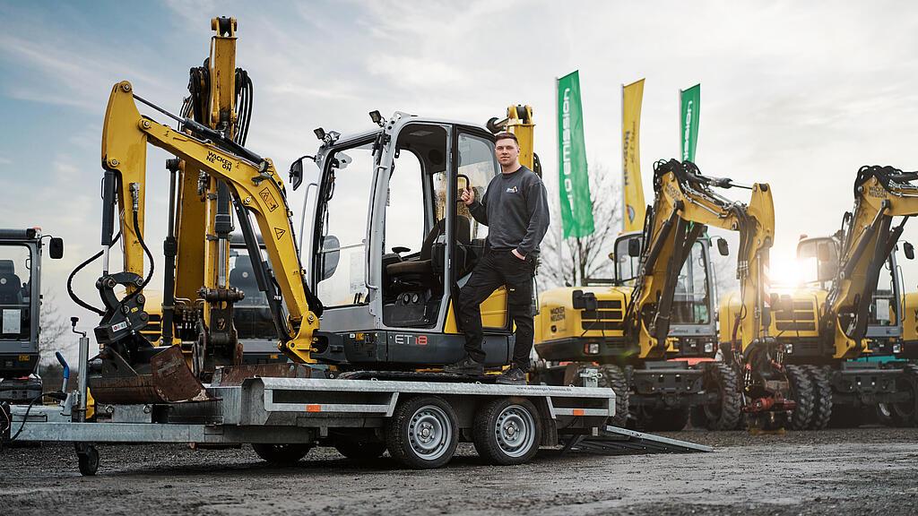 Ein Mann steht auf einem Anhänger neben einem Minibagger von Wacker Neuson auf einem Baumaschinenhof. Hinter ihm stehen weitere Bagger.