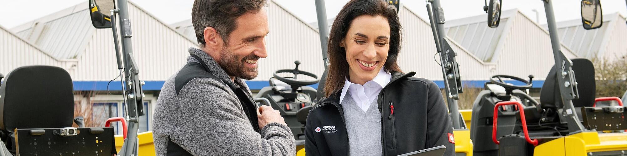 Smiling Wacker Neuson employee with tablet advising smiling customer in front of Wacker Neuson construction machines.