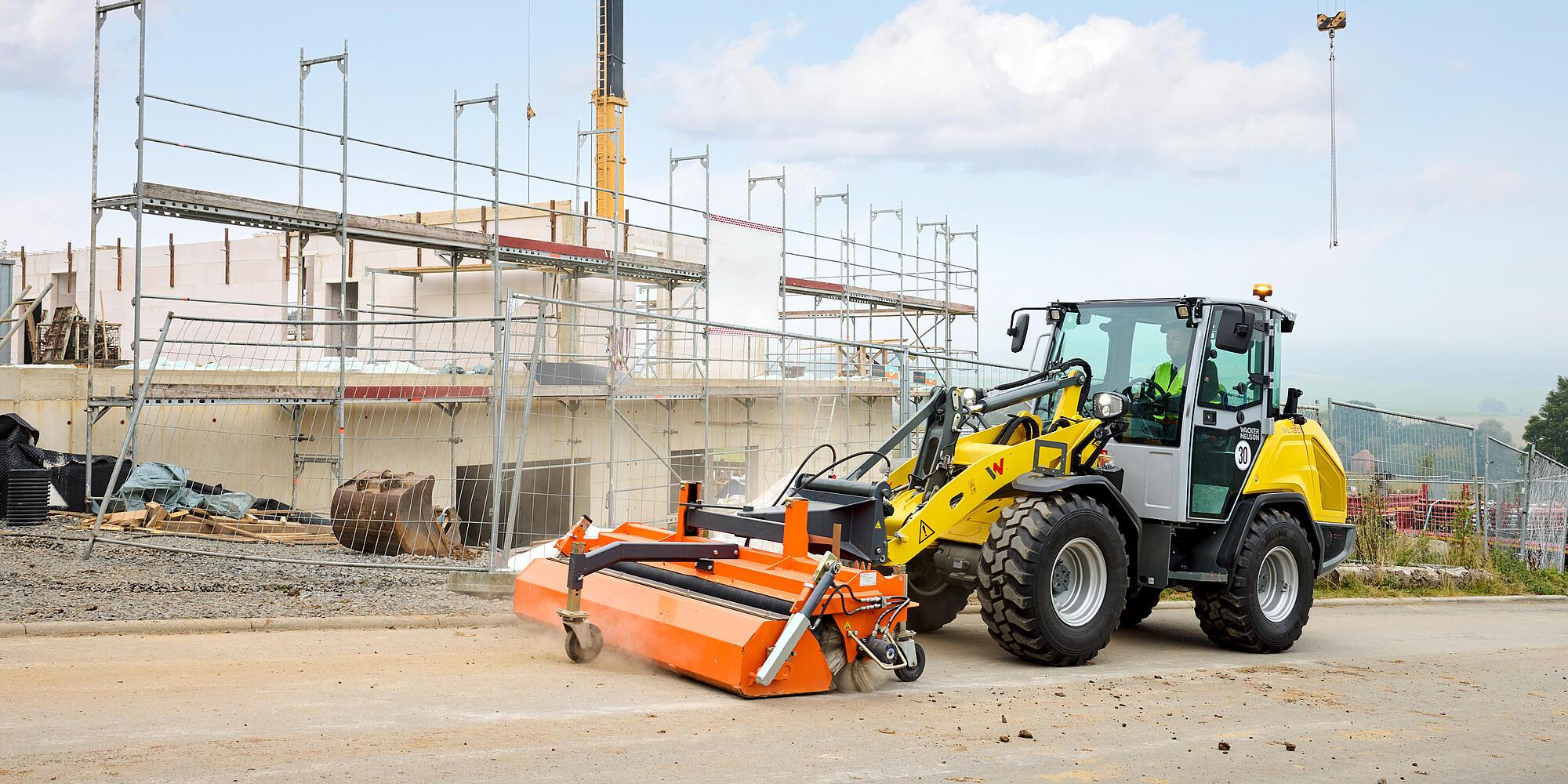 Wacker Neuson WL750 wheel loader equipped with a sweeper attachment cleaning a paved area in front of a building construction site with scaffolding and fencing in the background.